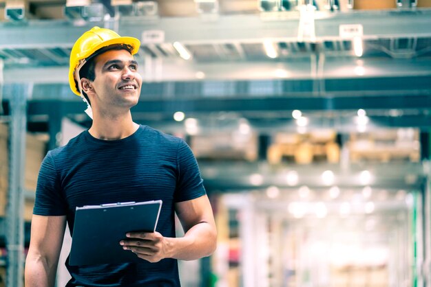 Uomo intelligente dell'ingegnere indiano che indossa un casco di sicurezza che esegue il controllo del segno di spunta e la gestione del prodotto in stock di cartone sullo sfondo del magazzino di fabbrica