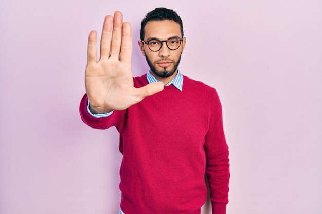 Uomo ispanico con barba che indossa camicia da lavoro e occhiali che smette di cantare con il palmo della mano. espressione di avvertimento con gesto negativo e serio sul viso.