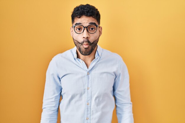 Uomo ispanico con barba in piedi su sfondo giallo che fa la faccia di pesce con labbra pazze e gesto comico espressione divertente