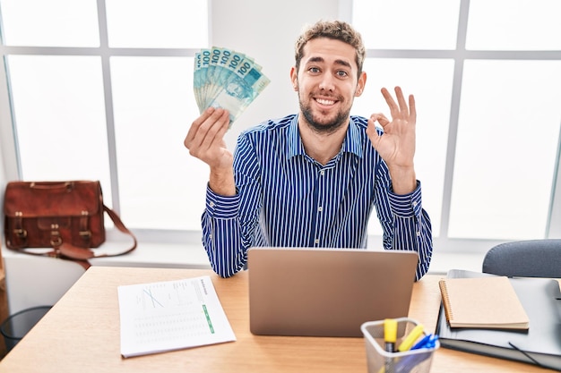 Uomo ispanico con la barba che lavora in ufficio con reals brasiliani che fanno segno ok con le dita sorridenti gesti amichevoli simbolo eccellente