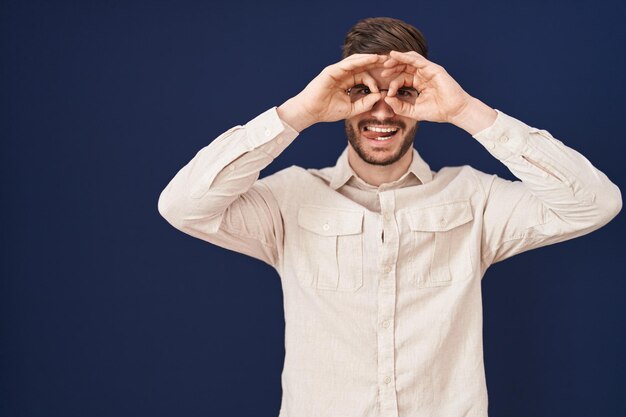 Uomo ispanico con la barba in piedi su sfondo blu facendo un gesto ok come un binocolo con la lingua fuori gli occhi guardando attraverso le dita espressione pazza