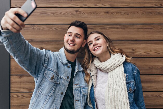 Uomo ispirato con la barba che fa selfie con la sua ragazza. Bellissima giovane donna con sciarpa nera in posa sulla parete in legno.