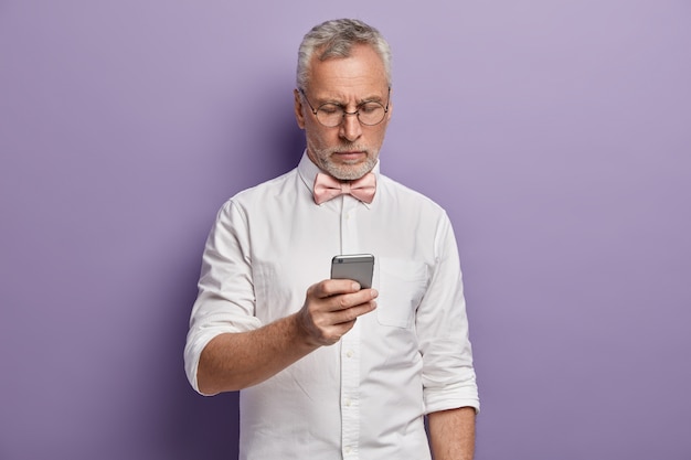 Uomo maggiore in camicia bianca e papillon rosa che tiene telefono