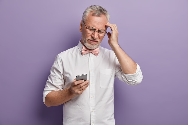 Uomo maggiore in camicia bianca e papillon rosa che tiene telefono