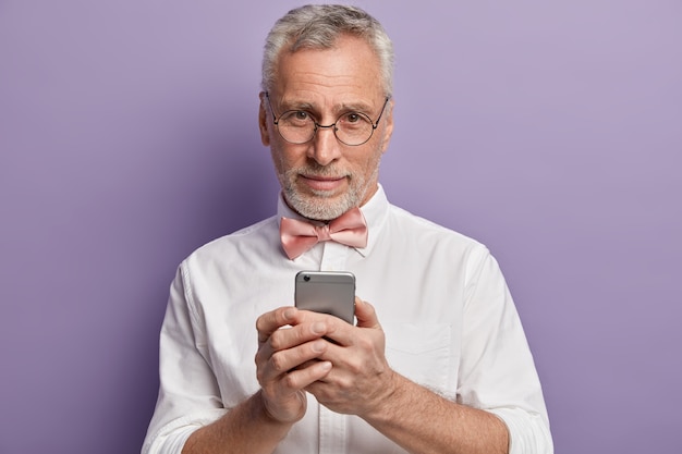 Uomo maggiore in camicia bianca e papillon rosa che tiene telefono