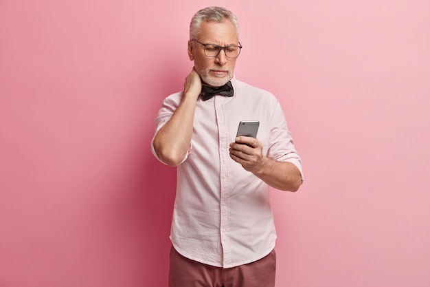 Uomo maggiore in camicia rosa e papillon nero che tiene il telefono