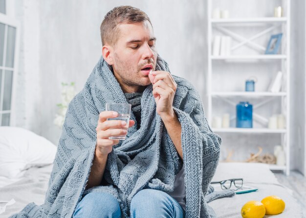 Uomo malato avvolto in sciarpa seduta sul letto a prendere la pillola con acqua
