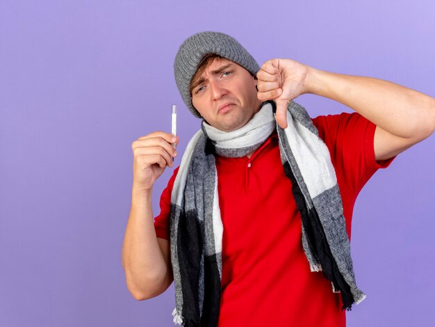 Uomo malato biondo bello giovane dispiaciuto che indossa cappello invernale e sciarpa che tiene il termometro guardando la parte anteriore che mostra il pollice verso il basso isolato sulla parete viola con lo spazio della copia