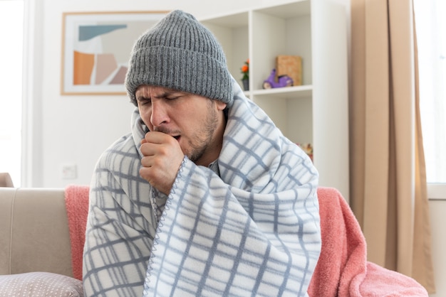 uomo malato dolorante con sciarpa intorno al collo che indossa cappello invernale avvolto in plaid tosse tenendo il pugno vicino alla bocca seduto sul divano in soggiorno