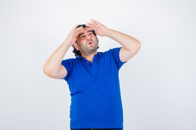 Uomo maturo che guarda lontano con la mano sopra la testa in maglietta blu, jeans e guardando sorpreso, vista frontale.