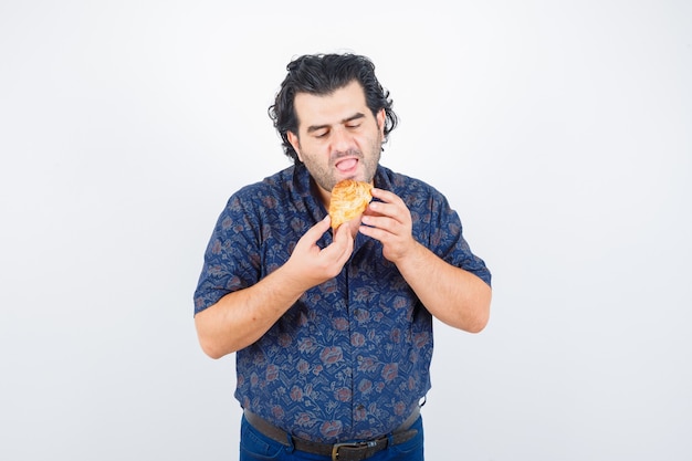 Uomo maturo che mangia prodotto di pasticceria in camicia e che sembra affamato, vista frontale.