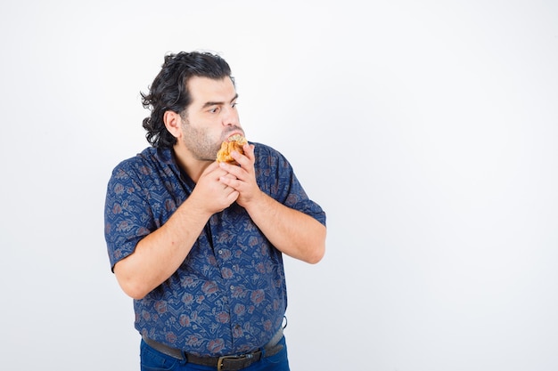 Uomo maturo che mangia prodotto di pasticceria mentre distoglie lo sguardo in camicia e sembra perplesso. vista frontale.