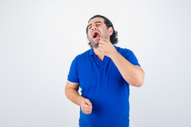 Uomo maturo che tiene la bocca spalancata, mettendo il dito indice vicino alla bocca in maglietta blu, jeans e sembra serio. vista frontale.