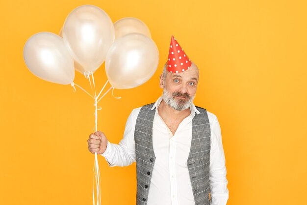 Uomo maturo con barba grigia che celebra anniversario. Ritratto di pensionato maschio bello con la barba lunga che tiene palloncini di elio divertendosi alla festa di compleanno.