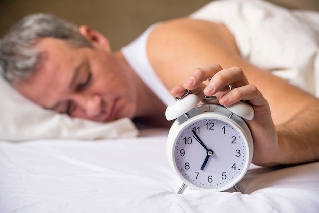 Uomo maturo dormire a letto con sveglia in primo piano alla camera da letto. L&#39;uomo esausto è svegliato da una sveglia nella sua camera da letto. Svegliato