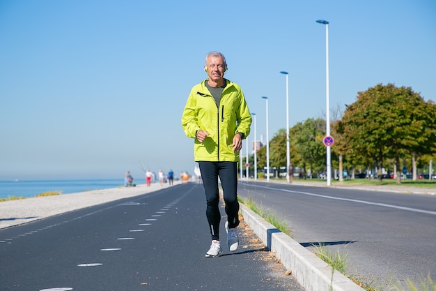 Uomo maturo eccitato felice in cuffie senza fili che pareggiano lungo la riva del fiume all'esterno. Pareggiatore senior formazione per la maratona. Vista frontale. Attività e concetto di età