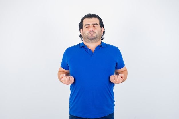 Uomo maturo meditando, tenendo gli occhi chiusi in maglietta blu, jeans e guardando calmo, vista frontale.