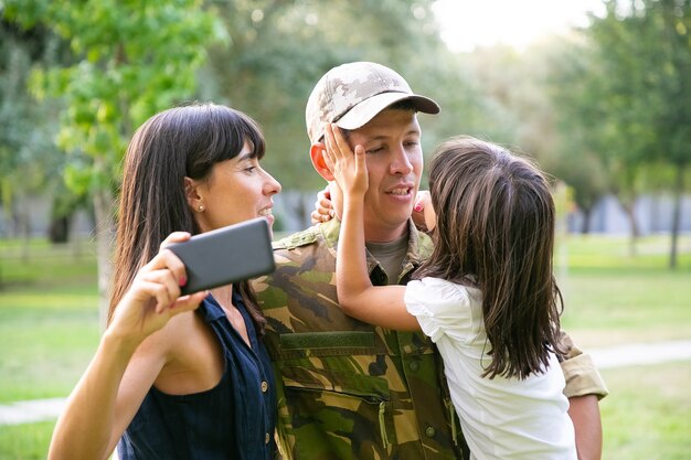 Uomo militare felice che gode del tempo insieme alla moglie e alla piccola figlia, prendendo selfie sul cellulare nel parco cittadino. Colpo medio. Ricongiungimento familiare o concetto di ritorno a casa