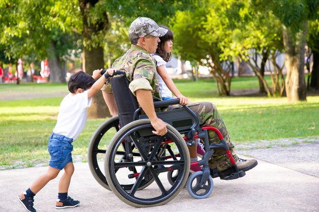 Uomo militare in pensione disabile che cammina con i bambini nel parco. Ragazza seduta sulle ginocchia di papà, ragazzo che spinge la sedia a rotelle. Veterano di guerra o concetto di disabilità