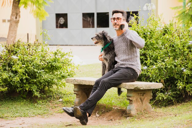 Uomo moderno che si siede nel parco con il suo cane che parla sul telefono cellulare