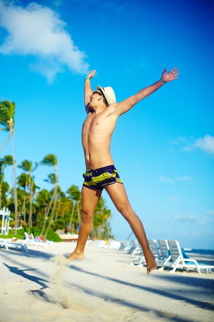 Uomo muscoloso bello felice in cappellino da sole sulla spiaggia che salta dietro il cielo blu dietro il cielo blu