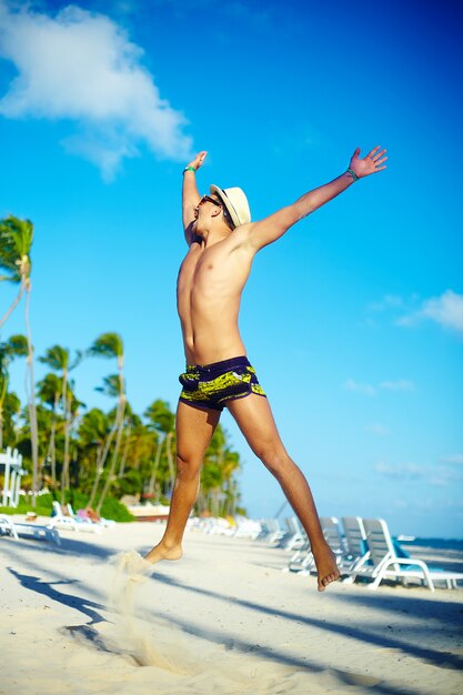 Uomo muscoloso bello felice in cappellino da sole sulla spiaggia che salta dietro il cielo blu dietro il cielo blu
