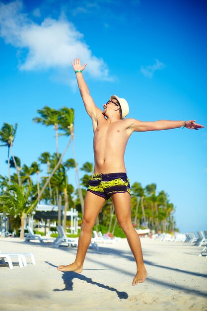 Uomo muscoloso bello felice in cappellino da sole sulla spiaggia che salta dietro il cielo blu dietro il cielo blu