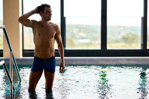 Uomo muscoloso in piedi in piscina e guardando lontano mentre trascorre un fine settimana al centro benessere Copia spazio
