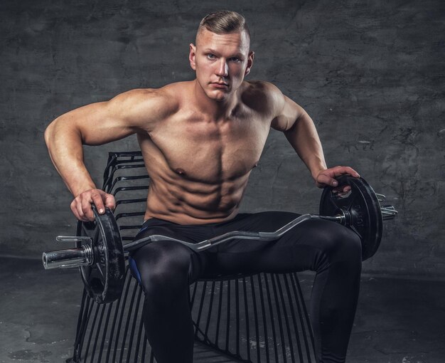Uomo muscoloso senza camicia con bilanciere si siede su una sedia nera.