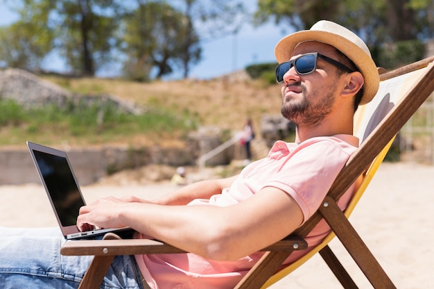 Uomo nella sedia di spiaggia che lavora al computer portatile