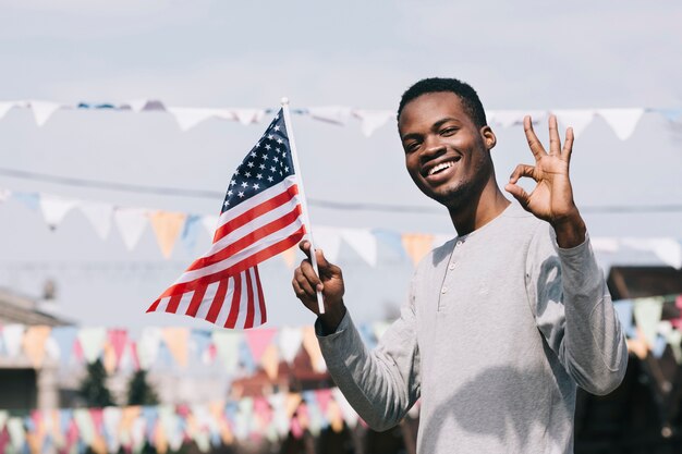 Uomo nero che tiene bandiera americana e che guarda l&#39;obbiettivo con gesto OK