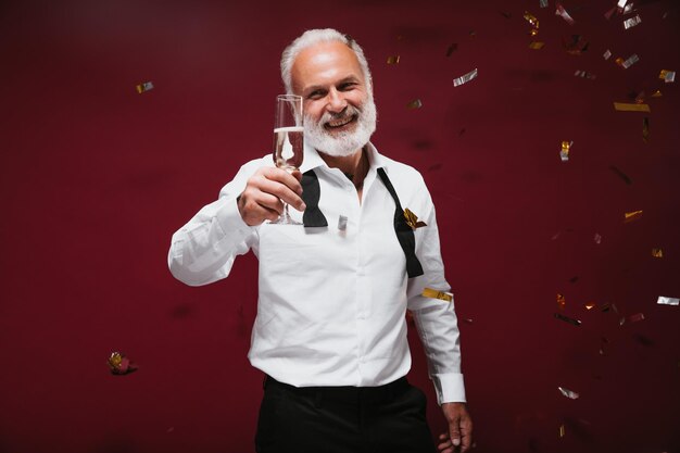 Uomo ottimista che ride e tiene un bicchiere di champagne Bel ragazzo barbuto in abito bianco in posa con vino su sfondo bordeaux