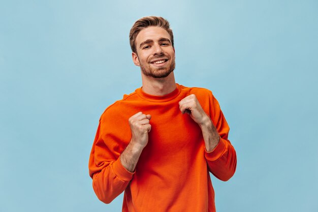 Uomo ottimista con barba rossa e bel sorriso in felpa arancione brillante che guarda la telecamera sul muro blu isolato