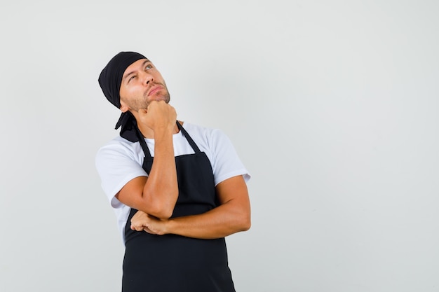 Uomo panettiere in t-shirt, grembiule che osserva in su con il mento appoggiato sulla mano e che sembra pensieroso