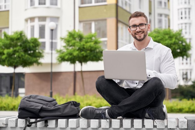 Uomo positivo che lavora su una panchina all'aperto in città utilizzando il computer portatile