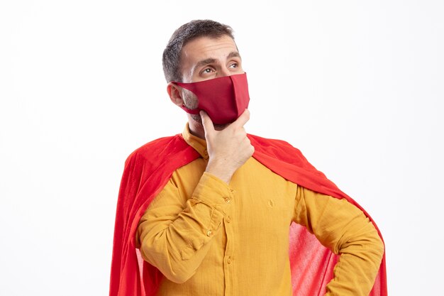 Uomo premuroso del supereroe con mantello rosso che indossa la maschera rossa mette la mano sul mento e guarda al lato isolato sul muro bianco