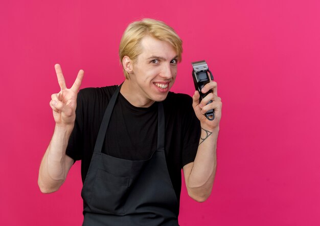 Uomo professionista del barbiere in grembiule che tiene trimmer sorridente felice e positivo che mostra il segno di v