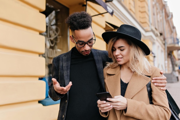 Uomo riccio divertente che guarda lo schermo del telefono e abbraccia una ragazza caucasica. Foto all'aperto di un ragazzo africano in abiti neri che cammina per la città con una magnifica donna bionda.