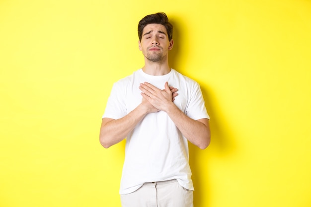 Uomo romantico e nostalgico che tiene le mani sul cuore, chiudi gli occhi e ricorda qualcosa, in piedi su sfondo giallo.