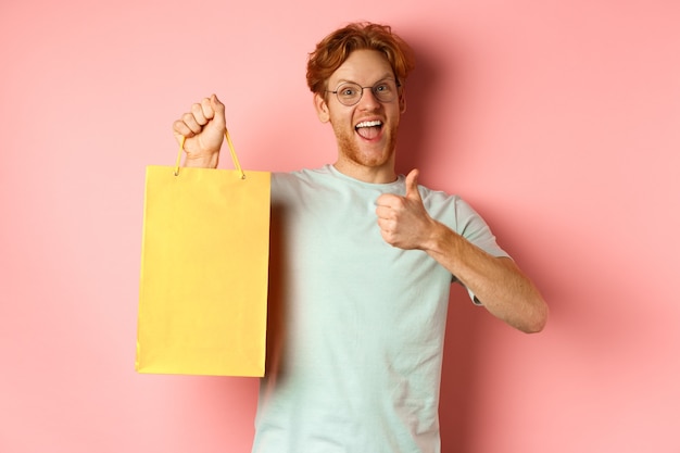 Uomo rosso allegro in t-shirt e occhiali che punta il dito contro la borsa della spesa, mostrando il negozio con sconti, in piedi su sfondo rosa