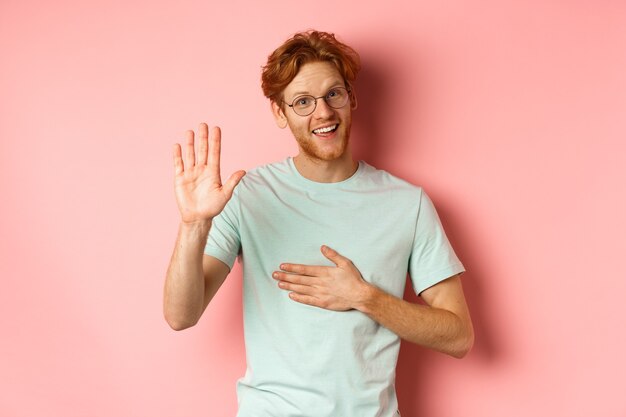 Uomo rosso amichevole che è onesto, tenendo la mano sul cuore e il braccio alzato in alto per giurare o fare promesse, sorridere alla telecamera, dicendo la verità su sfondo rosa.