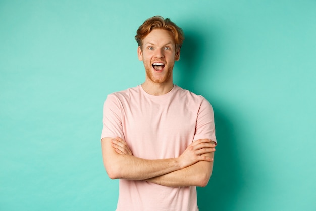 Uomo rosso entusiasta in t-shirt che controlla promo interessante, braccia incrociate sul petto e guardando la telecamera con stupore, in piedi su sfondo turchese