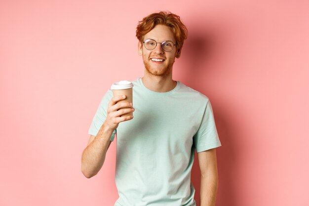 Uomo rosso felice in bicchieri e t-shirt bere caffè e sorridere, godersi la pausa pranzo, tenendo tazza da asporto, in piedi su sfondo rosa.