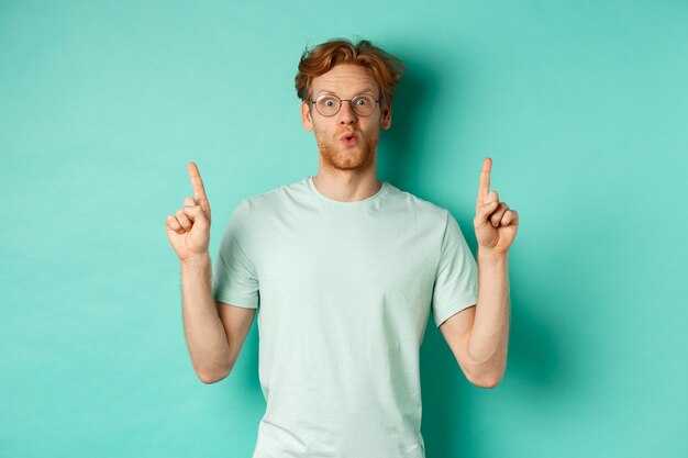 Uomo rosso impressionato in occhiali e t-shirt, controllando l'offerta promozionale, puntando le dita verso lo spazio della copia, fissando la telecamera stupito, in piedi su sfondo turchese
