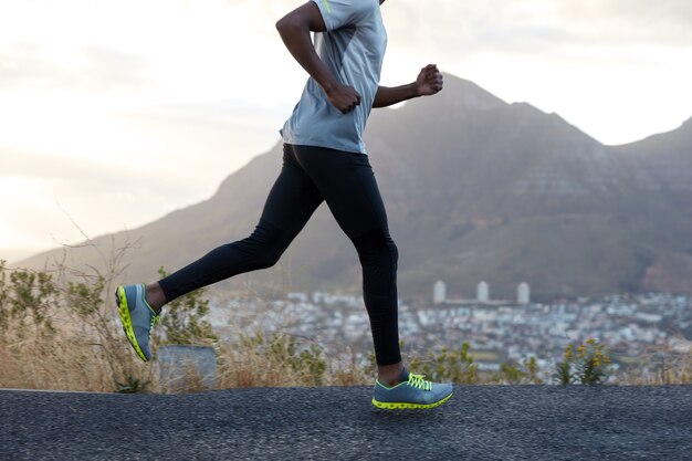 Uomo sano dalla pelle scura in azione, corre lungo la strada vicino alle montagne, indossa scarpe da ginnastica comode, abiti casual, ha un corpo sportivo. Atleta maschio veloce pone contro il cielo. Competizione di corse