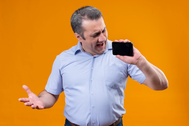 Uomo scontento e arrabbiato in camicia a righe blu guardando il suo telefono cellulare in piedi