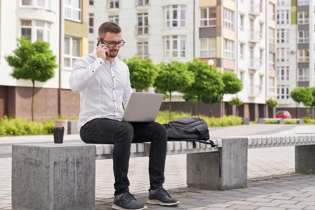 Uomo seduto su una panchina che lavora al computer portatile parlando al telefono