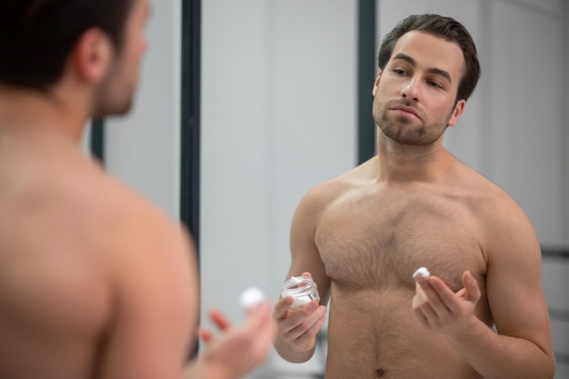 Uomo seminudo in piedi vicino allo specchio e applicando schiuma da barba sul viso