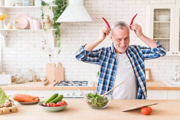 Uomo senior che tiene peperoncino rosso vicino alla testa come corna del diavolo rosso in cucina