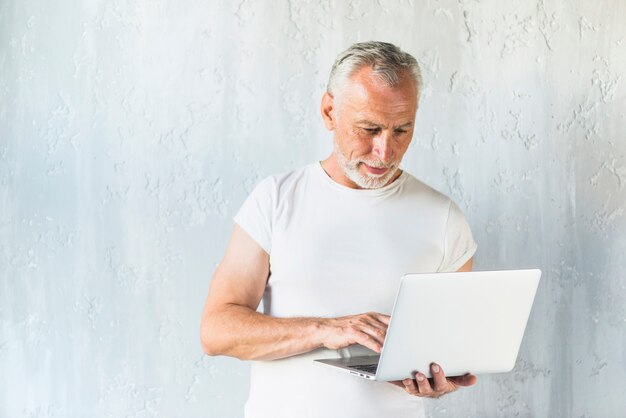 Uomo senior che utilizza computer portatile che sta davanti al muro di cemento